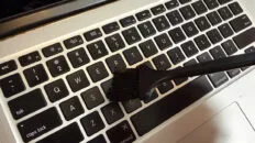 Laptop held at an angle while a soft brush removes debris between keys