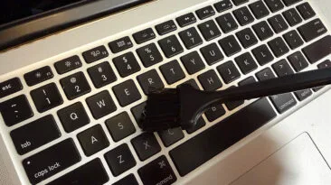 Laptop held at an angle while a soft brush removes debris between keys